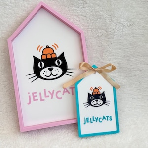 Puede incluir: Un marco rosa con forma de casa que contiene un letrero blanco con un gato negro con sombrero naranja y el texto JELLYCAT en rosa, junto a una etiqueta turquesa m&aacute;s peque&ntilde;a con un gr&aacute;fico de gato, el texto JELLYCATS y un lazo beige.