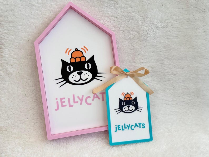 Puede incluir: Un marco rosa con forma de casa que contiene un letrero blanco con un gato negro con sombrero naranja y el texto JELLYCAT en rosa, junto a una etiqueta turquesa m&aacute;s peque&ntilde;a con un gr&aacute;fico de gato, el texto JELLYCATS y un lazo beige.
