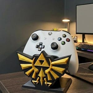 Peut inclure: Une manette Xbox blanche repose sur un support décoratif noir et doré. Le support présente un emblème doré avec une base noire. La manette a des boutons noirs et des boutons d'action colorés.