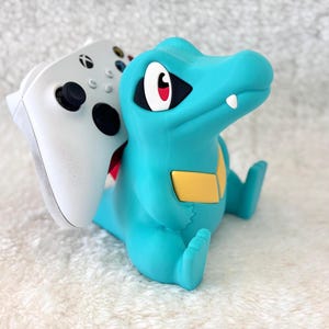 May include: A teal-colored, cartoon-style figure of a crocodile holding a white Xbox controller. The crocodile has a yellow belly patch, red eyes, and a white tooth. The controller is white with black buttons and joysticks.