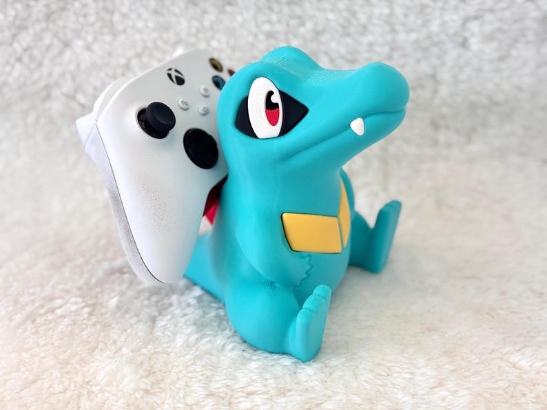 May include: A teal-colored, cartoon-style figure of a crocodile holding a white Xbox controller. The crocodile has a yellow belly patch, red eyes, and a white tooth. The controller is white with black buttons and joysticks.