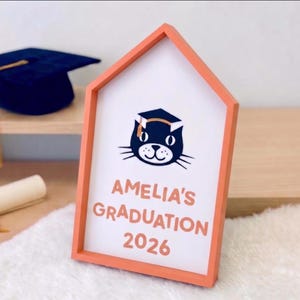 May include: A graduation-themed sign in a house-shaped frame. The sign features a cat wearing a graduation cap, the name "Amelia's", and the year "2026" in orange lettering. A navy blue graduation cap with a gold tassel is visible in the background.