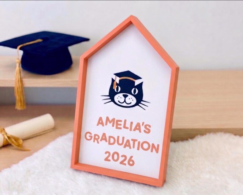 May include: A graduation-themed sign in a house-shaped frame. The sign features a cat wearing a graduation cap, the name "Amelia's", and the year "2026" in orange lettering. A navy blue graduation cap with a gold tassel is visible in the background.