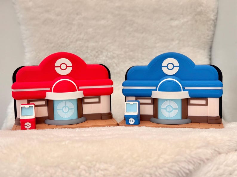 May include: Two colorful, stylized building models. One is red, the other blue, each with a white and red logo on top. They have a white archway and a light blue door. A matching red and blue device sits in front of each building.