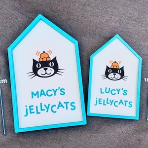 May include: Two house-shaped signs with a teal frame. The signs feature a black cat wearing a pumpkin hat and the text "MACY'S JELLYCATS" and "LUCY'S JELLYCATS". The larger sign is 26.2 cm tall, and the smaller sign is 18.8 cm tall.