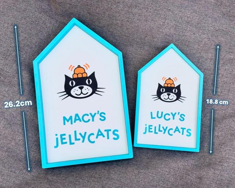 May include: Two house-shaped signs with a teal frame. The signs feature a black cat wearing a pumpkin hat and the text "MACY'S JELLYCATS" and "LUCY'S JELLYCATS". The larger sign is 26.2 cm tall, and the smaller sign is 18.8 cm tall.