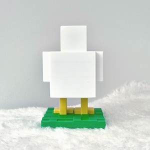 May include: A white, blocky chicken figurine with yellow legs and a green base. The chicken has a minimalist, pixelated design, reminiscent of video game graphics. The figurine is set against a soft, white background.