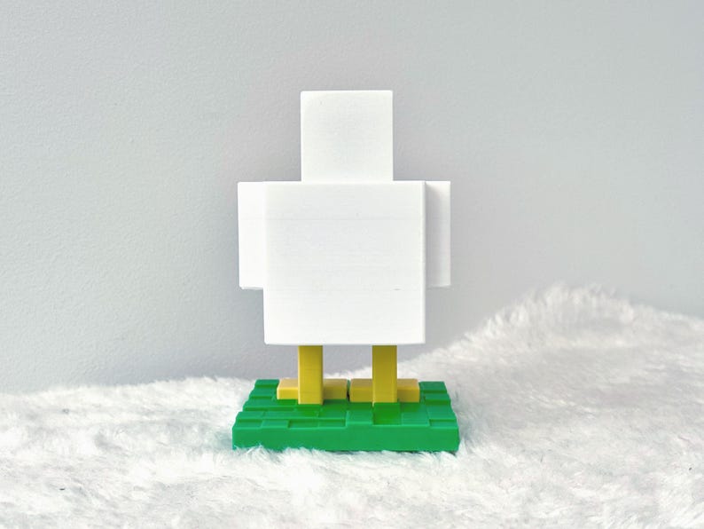May include: A white, blocky chicken figurine with yellow legs and a green base. The chicken has a minimalist, pixelated design, reminiscent of video game graphics. The figurine is set against a soft, white background.