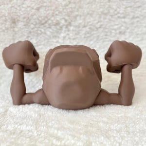 May include: A brown action figure torso with arms raised in a flexing pose. The figure has a detailed design, with defined muscles and a smooth, matte finish. The arms are bent at the elbows, with clenched fists.