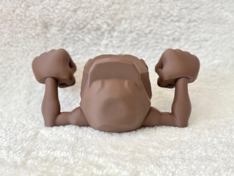 May include: A brown action figure torso with arms raised in a flexing pose. The figure has a detailed design, with defined muscles and a smooth, matte finish. The arms are bent at the elbows, with clenched fists.