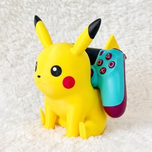 May include: A yellow Pikachu character figurine serving as a controller stand. The figure has black-tipped ears and red cheeks. A teal and purple video game controller with white symbols is resting securely against the back of the yellow figure.