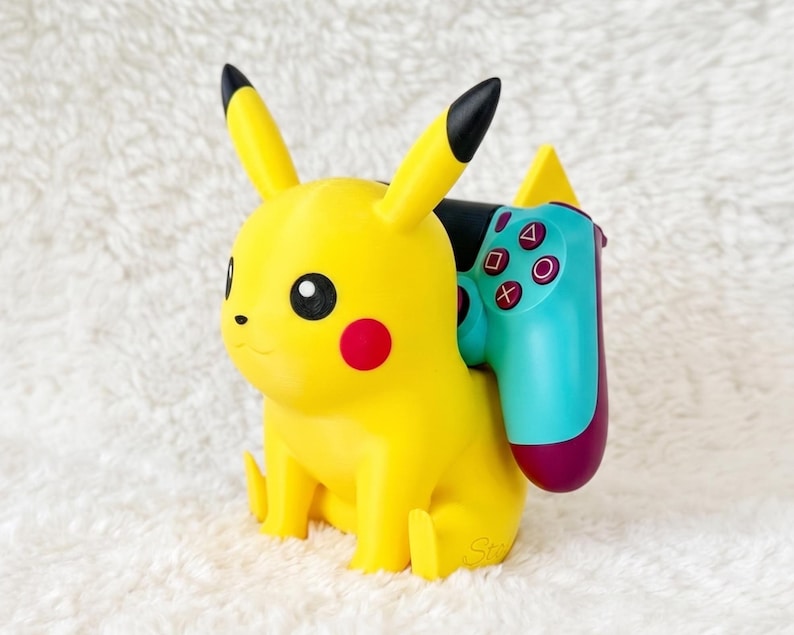 May include: A yellow Pikachu character figurine serving as a controller stand. The figure has black-tipped ears and red cheeks. A teal and purple video game controller with white symbols is resting securely against the back of the yellow figure.