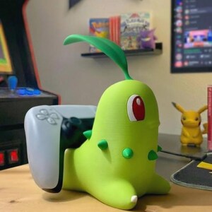 May include: A green Chikorita-shaped controller holder with a white PlayStation 5 controller. The Chikorita figure has a red eye, green spikes, and a leaf on its head. A yellow Pikachu figure is visible in the background.