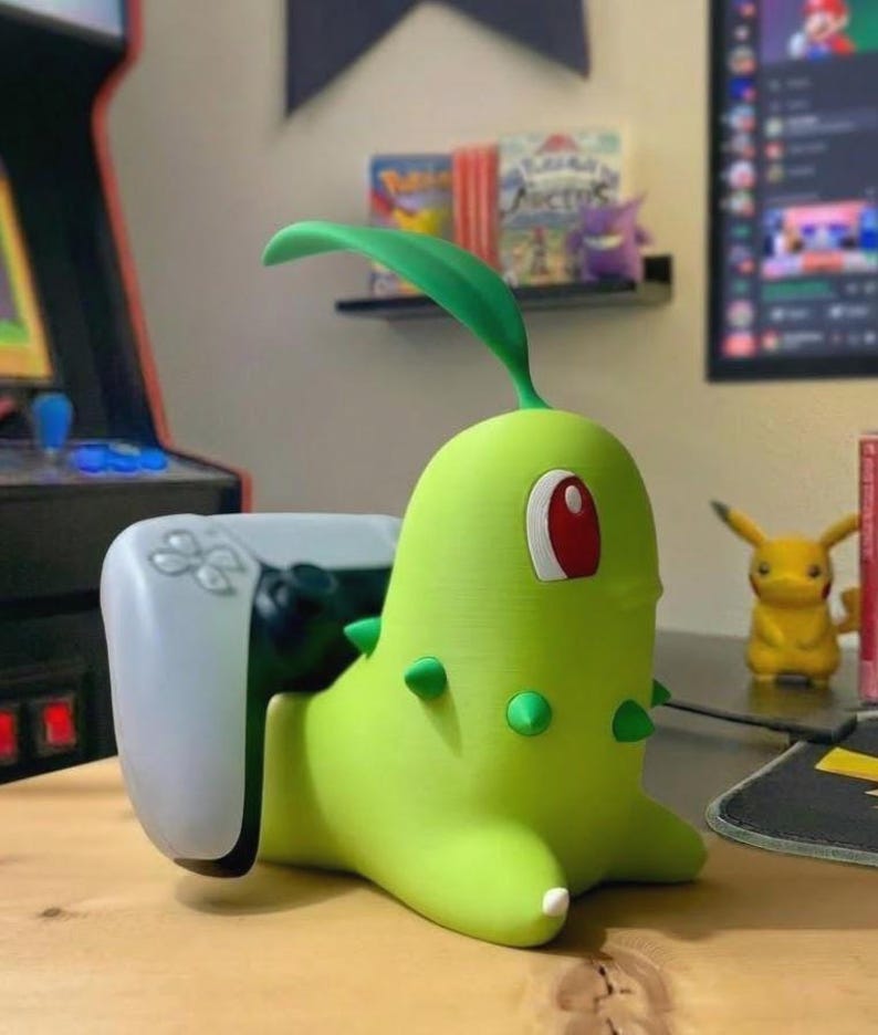 May include: A green Chikorita-shaped controller holder with a white PlayStation 5 controller. The Chikorita figure has a red eye, green spikes, and a leaf on its head. A yellow Pikachu figure is visible in the background.