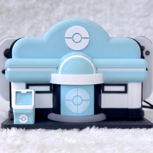 May include: A light blue and white gaming console dock shaped like a building with a circular emblem on the front. The structure features a central entrance, a small side kiosk, and a dark base, designed to hold a handheld gaming device securely.