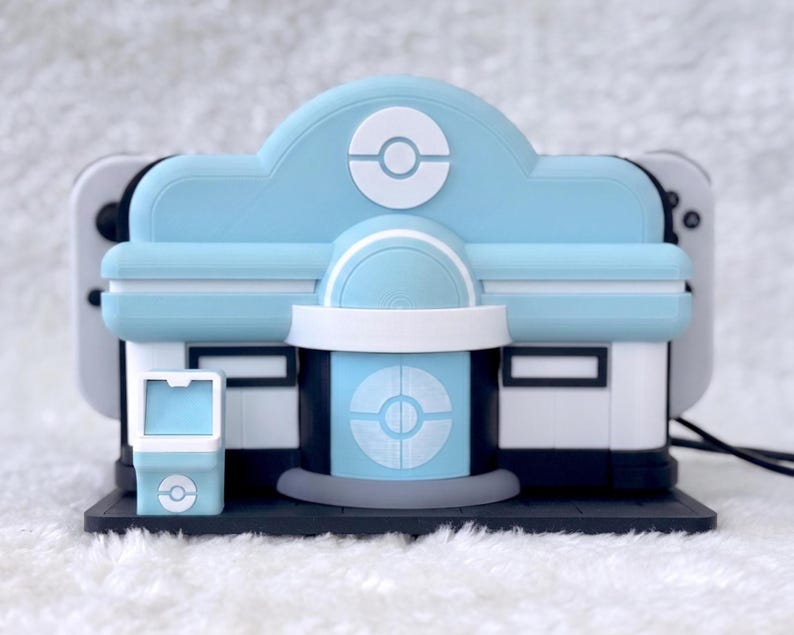 May include: A light blue and white gaming console dock shaped like a building with a circular emblem on the front. The structure features a central entrance, a small side kiosk, and a dark base, designed to hold a handheld gaming device securely.