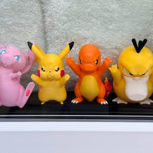 May include: Four small plastic character figurines standing in a row. From left to right: a pink creature with a long tail, a yellow creature with long ears and red cheeks, an orange creature with a flame tail, and a yellow creature with black hair.