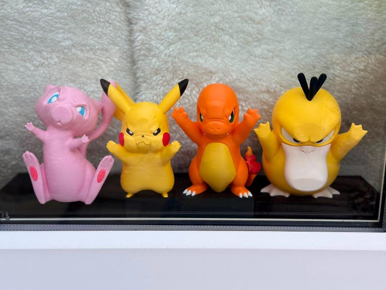 May include: Four small plastic character figurines standing in a row. From left to right: a pink creature with a long tail, a yellow creature with long ears and red cheeks, an orange creature with a flame tail, and a yellow creature with black hair.