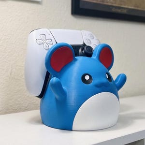 May include: A blue and red cartoon character-shaped controller stand. The stand is holding a white game controller. The character has large red ears and a smiling face. The stand is designed to hold a game controller.