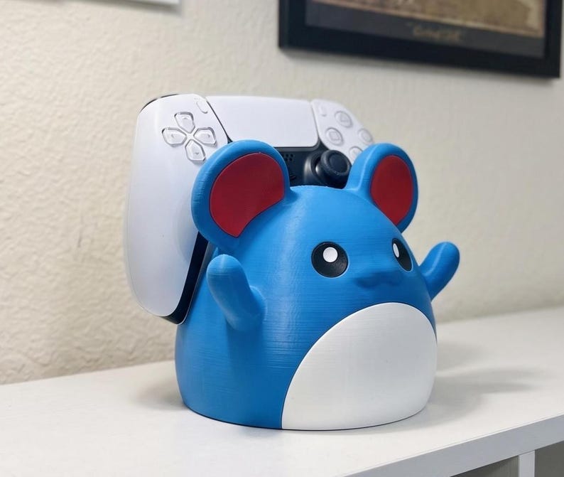 May include: A blue and red cartoon character-shaped controller stand. The stand is holding a white game controller. The character has large red ears and a smiling face. The stand is designed to hold a game controller.