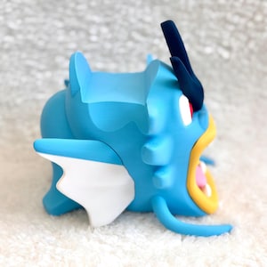 May include: A blue figurine of a sea creature with a large open mouth, yellow lips, and white teeth. It features a dark blue crest on its head, white side fins, and a smooth matte finish. The creature has a prominent red eye and a curved body.