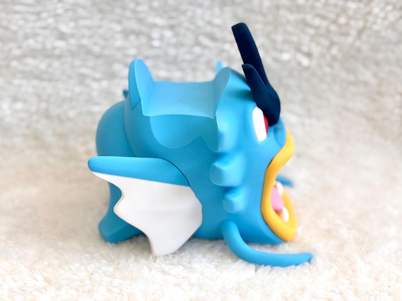 May include: A blue figurine of a sea creature with a large open mouth, yellow lips, and white teeth. It features a dark blue crest on its head, white side fins, and a smooth matte finish. The creature has a prominent red eye and a curved body.