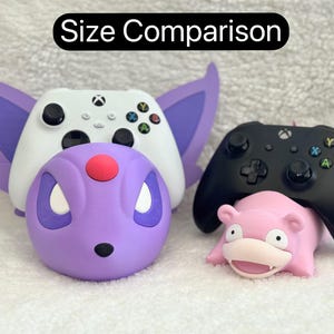 Puede incluir: Un mando de Xbox blanco sobre un soporte morado con forma de criatura y orejas grandes, junto a un mando de Xbox negro sobre un soporte rosa con forma de criatura. El texto "Size Comparison" aparece en la parte superior.