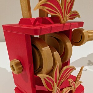 May include: A red and gold decorative object with a geometric design. The object features a series of gold-colored wheels and decorative leaf-like elements. The structure has a boxy shape with a handle and a knob.