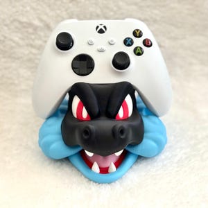 May include: A white Xbox wireless controller resting on a black and light blue character-shaped stand. The stand features a black snout, red eyes, white teeth, and a light blue jawline, designed to hold the gaming controller securely in place.