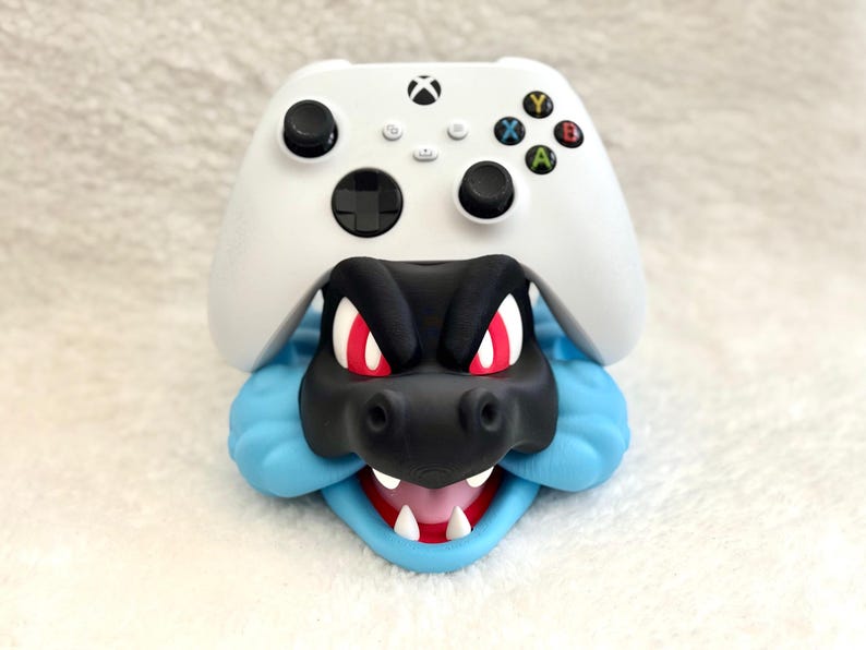 May include: A white Xbox wireless controller resting on a black and light blue character-shaped stand. The stand features a black snout, red eyes, white teeth, and a light blue jawline, designed to hold the gaming controller securely in place.