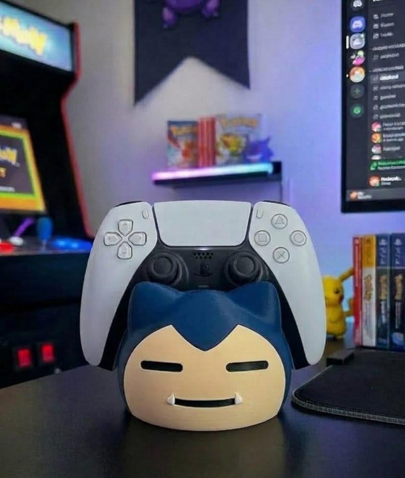 May include: A white and black PlayStation 5 controller sits on a blue and tan Snorlax-shaped stand. The stand is on a black surface. The background includes a shelf with video games and a Pikachu figure.