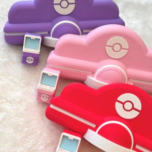 May include: Three colorful, stylized cloud-shaped accessories in purple, pink, and red, each with a white circular emblem. Accompanying each cloud is a matching small, rectangular device with a light blue screen and a similar emblem.