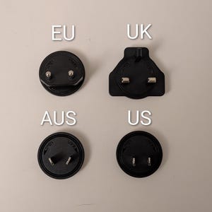 May include: Four black electrical plug adapters, labeled EU, UK, AUS, and US, are arranged on a light beige surface. The EU and AUS adapters are circular, while the UK adapter is rectangular. The US adapter is also circular.