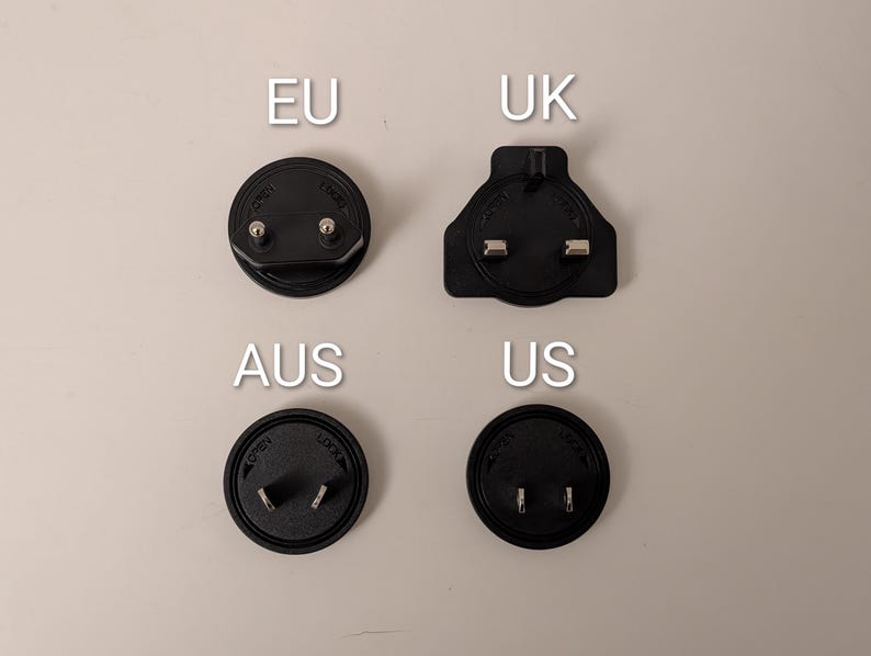 May include: Four black electrical plug adapters, labeled EU, UK, AUS, and US, are arranged on a light beige surface. The EU and AUS adapters are circular, while the UK adapter is rectangular. The US adapter is also circular.