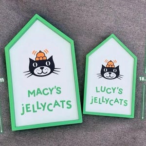 May include: Two house-shaped signs with green frames. The larger sign is 26.2 cm tall and reads "MACY'S JELLYCATS" below a black cat illustration wearing a hat. The smaller sign is 18.8 cm tall and reads "LUCY'S JELLYCATS".
