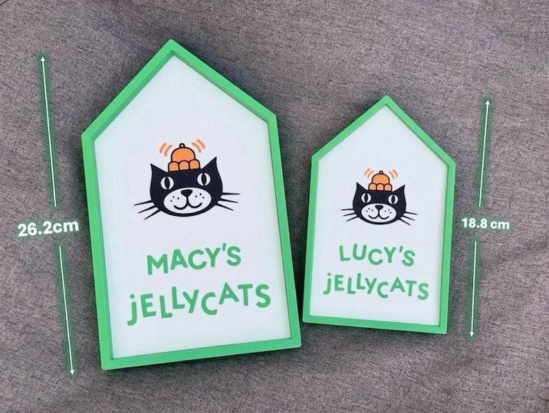 May include: Two house-shaped signs with green frames. The larger sign is 26.2 cm tall and reads "MACY'S JELLYCATS" below a black cat illustration wearing a hat. The smaller sign is 18.8 cm tall and reads "LUCY'S JELLYCATS".