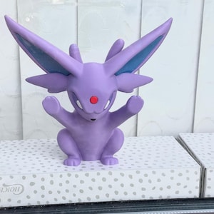 Puede incluir: Una figura de Espeon de color lavanda con grandes orejas puntiagudas, una gema roja en la frente y una cola dividida. La criatura est&aacute; representada en posici&oacute;n sentada con los brazos levantados, con detalles azules en el interior de las orejas.