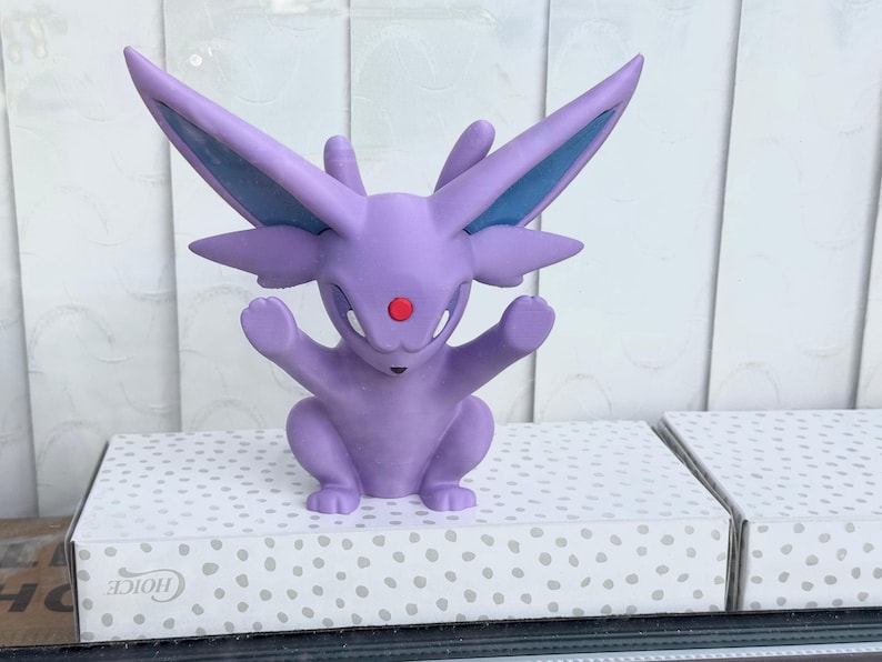 Puede incluir: Una figura de Espeon de color lavanda con grandes orejas puntiagudas, una gema roja en la frente y una cola dividida. La criatura est&aacute; representada en posici&oacute;n sentada con los brazos levantados, con detalles azules en el interior de las orejas.