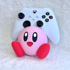 May include: A white wireless video game controller with black joysticks and coloured action buttons positioned behind a round, pink Kirby character figure with red feet, large oval eyes, and a small open mouth, all set against a white textured background.