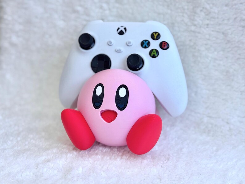 May include: A white wireless video game controller with black joysticks and coloured action buttons positioned behind a round, pink Kirby character figure with red feet, large oval eyes, and a small open mouth, all set against a white textured background.