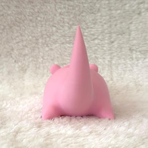 Pode incluir: Uma figura de rinoceronte rosa com um chifre proeminente e orelhas pequenas. O rinoceronte &eacute; retratado em posi&ccedil;&atilde;o sentada, com as pernas dobradas sob o corpo. A figura &eacute; feita de um material liso e est&aacute; sobre um fundo branco texturizado.