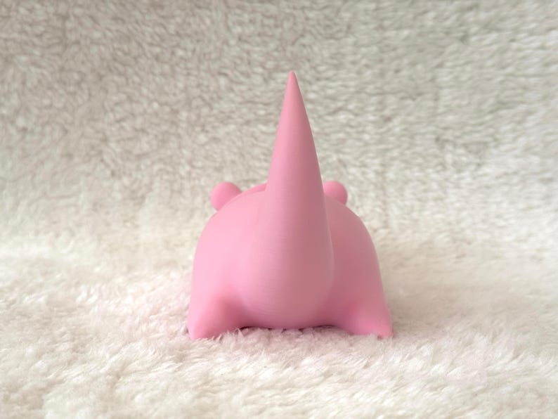 Pode incluir: Uma figura de rinoceronte rosa com um chifre proeminente e orelhas pequenas. O rinoceronte &eacute; retratado em posi&ccedil;&atilde;o sentada, com as pernas dobradas sob o corpo. A figura &eacute; feita de um material liso e est&aacute; sobre um fundo branco texturizado.