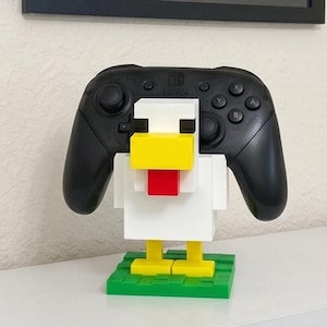May include: A black Nintendo Switch Pro controller rests in a white, yellow, and red chicken-shaped holder made of interlocking blocks. The holder sits on a green base, creating a playful and functional accessory.