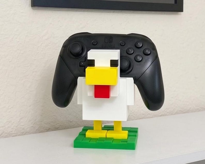 May include: A black Nintendo Switch Pro controller rests in a white, yellow, and red chicken-shaped holder made of interlocking blocks. The holder sits on a green base, creating a playful and functional accessory.