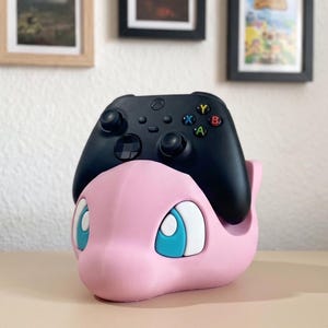 May include: A pink, 3D-printed controller holder shaped like a cartoon character, with blue and white eyes. A black Xbox controller rests on top. The holder is on a light brown surface, with framed pictures in the background.