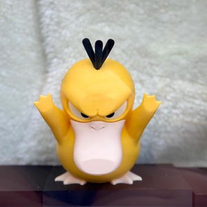 May include: A yellow, duck-like creature figurine with a large, pale pink beak, three black hair-like tufts on its head, and a stern facial expression. The figure has its arms raised upward and is standing on two pale pink webbed feet.