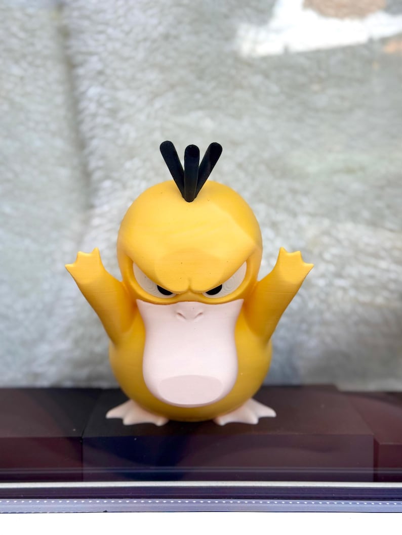 May include: A yellow, duck-like creature figurine with a large, pale pink beak, three black hair-like tufts on its head, and a stern facial expression. The figure has its arms raised upward and is standing on two pale pink webbed feet.