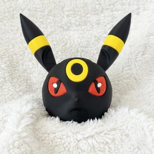 May include: A black figurine head featuring two long pointed ears with yellow bands, red eyes with white pupils, and a yellow circular marking on the forehead. The character has a smooth matte finish and a neutral expression with a small nose and mouth.