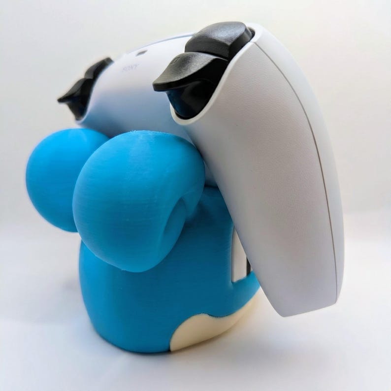 May include: A blue and white gaming controller stand shaped like a cartoon character. The stand has two blue spheres on the sides and a white controller resting on top. The stand is designed to hold a gaming controller.