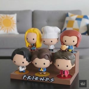 May include: A collection of six cartoon character figurines from the TV show "Friends" displayed on a brown stand. The figurines have large heads and small bodies, with the show's logo "F.R.I.E.N.D.S" printed on the base.